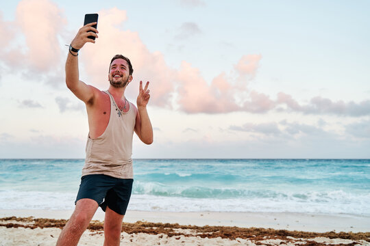 portrait caucasian man with a cell phone in his hands taking a selfie picture and making bunnies at sunset at the sea of cancun
