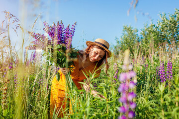 Young woman holding bouquet of lupin flowers walking in summer meadow. Stylish girl picking purple...