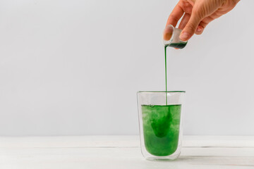 Woman hand pouring liquid chlorophyll in a glass cup. White background - obrazy, fototapety, plakaty