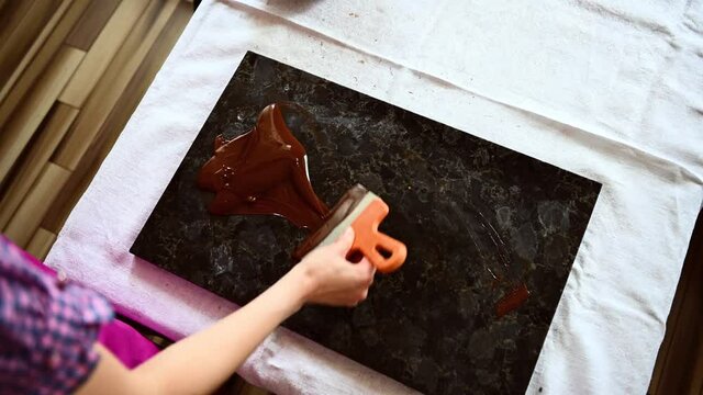 Top view video with a chocolatier tempering melted liquid dark chocolate on a marble table using a cake scraper to cool it and prepare delicious chocolates. World Chocolate Day. Cooking video lessons.