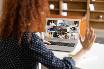 Close-up of curly woman waving at the laptop screen with people profiles, using an application for distance video communication with coworkers, webinar participants, meeting online in pandemic