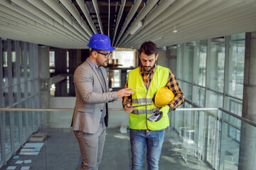Architect and construction worker standing in building in construction process and looking at blueprints on tablet.