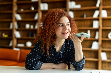 Smiling young redhead woman using convert voice into text mobile app, holding smartphone and talking into a mic. Charming curly girl recording and sending voice message, writing sms by voice - Powered by Adobe