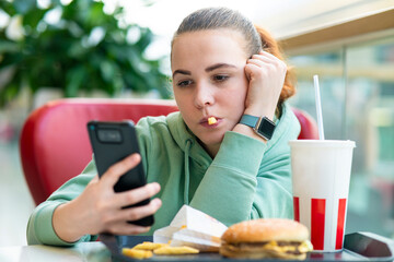 Young beautiful woman, bored girl eating fast junk food: burger, hamburger, potato French fries and...