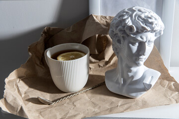 A white corrugated cup with tea and lemon, with a plaster head sculpture of Apollo and a...