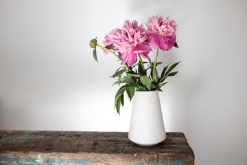 Red, pink peonies in a white vase on an old bench against a white wall