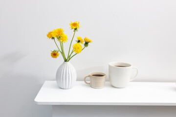 A bouquet of dandelions in a white fluted vase and two cups of different sizes with coffee on a white table.