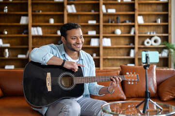Handsome attractive indian guy making video blog about musical instruments, sitting on the couch,...