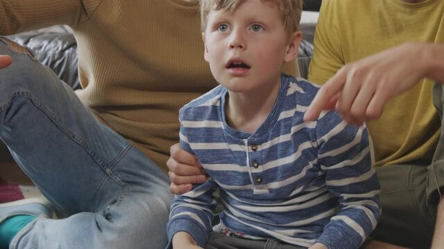 Slowmo tilt up shot of happy little boy sitting on rug and playing video game while his gay fathers watching He is winning and celebrating