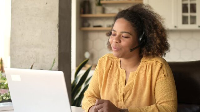 Cheerful attractive African-American woman wearing wireless headset using a laptop for video connection, talking online, a saleswoman takes a call working remotely from home office