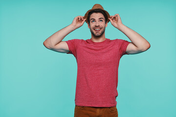 Fototapeta premium Handsome young man adjusting his fedora hat and smiling while standing against blue background