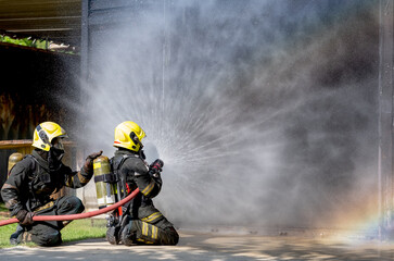 Two firefighters sit on floor and spray of water with curtain shape and rainbow reflex occur on water aerosol in front of container containing fire inside.