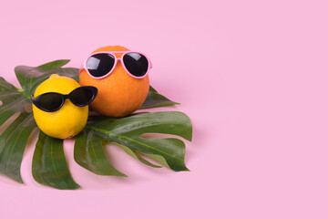 Orange and lemon fruit hipster in sunglasses on monstera leaf on pink background. Minimal travel concept, summer stylish tropical fruit.