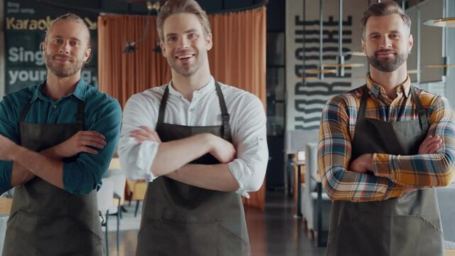 Handsome male waiters in aprons keeping arms crossed and looking at camera 
