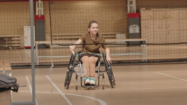 Long shot of fifteen-year-old Caucasian girl in wheelchair hitting shuttlecocks with badminton racket in indoor court