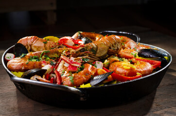 Paella traditional Spanish dish, served in the paella pan on a rustic wooden table highlighting the ingredients, shrimp, lobster, mussels, rice, red pepper and spices