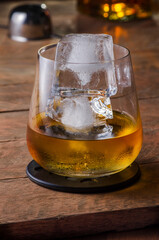 glass of whiskey with two ice cubes on a rustic wooden table with a bottle and a dispenser in the background