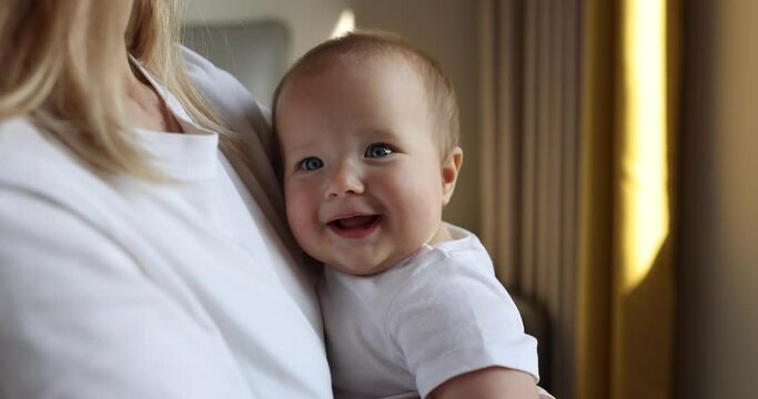 Young caucasian mother and her baby daughter hugging and playing at home in bedroom. Little kid seven months old spending time together with mom arms at home near window, slow motion.