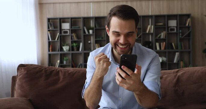 On-line bet, internet auction winner celebrate victory showing thumbs up. Millennial man sit on sofa holding smart phone check e-mail app read unbelievable news, got commercial offer feels overjoyed