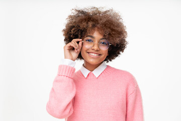 Smiling african teen girl in pink sweater holding eyeglasses with hand, looking at camera isolated on gray background