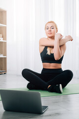 Stretching exercise. Sportive woman. Online fitness. Home gym. Athletic lady in black sportswear doing warming workout for hands looking laptop sitting sport mat in light room interior.