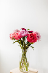 A bright image of a bouquet of flowers in a clear vase against a white wall. The peony flowers are cheerful in colors of pink, red, magenta, and violet.
