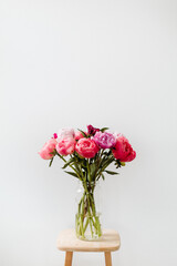 Naklejka na ściany i meble A bright image of a bouquet of flowers in a clear vase against a white wall. The peony flowers are cheerful in colors of pink, red, magenta, and violet.