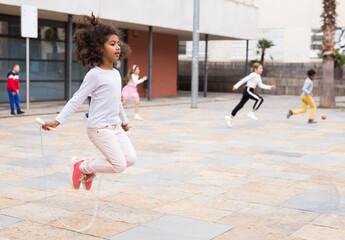 Cute tween African American girl skipping rope in schoolyard during break between lessons - obrazy, fototapety, plakaty