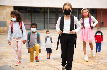 Portrait of positive schoolgirl in mask standing near school, children on background