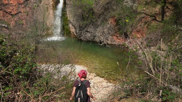 Young woman in dirty clothes and with backpack has finally found waterfall in forest while hiking, and hurries to it along the path, throwing off bag and rejoicing at discovery.