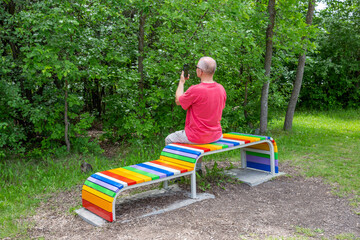 caucasian middle aged male sitting on a rainbow colored bench in the summer.