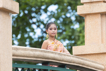 An Asian schoolgirl dressed in traditional Thai clothes stands at the railing of a bridge in a park.