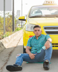 Portrait of a young Latino man sitting in front of a yellow taxi. Yellow car in the middle of the...