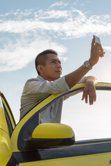 Latino man using his cell phone outside his yellow taxi. Hispanic driver in a gray shirt, taking a photo outside the car. Video call.