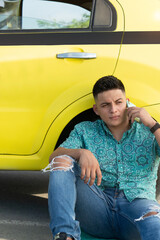 Latino man talking on the cell phone, sitting next to a yellow car. Hispanic man dressed casually...