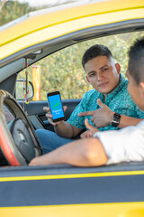Latino man showing the cell phone to the driver of the vehicle. Young man paying electronically to the yellow taxi driver.
