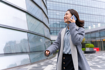 Young beautiful girl walking outside office building using mobile phone and happily calling customers
