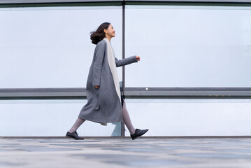 full length side view of Young stylish Chinese businesswoman walking outside modern office building...