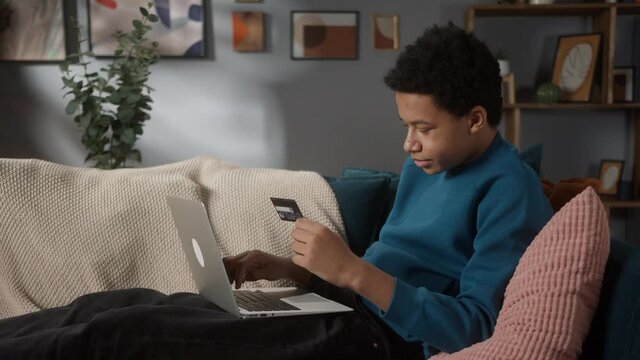 Black teenager man using credit card, entering pincode, african american male doing shopping online on laptop during coronavirus lockdown, high school student typing. 