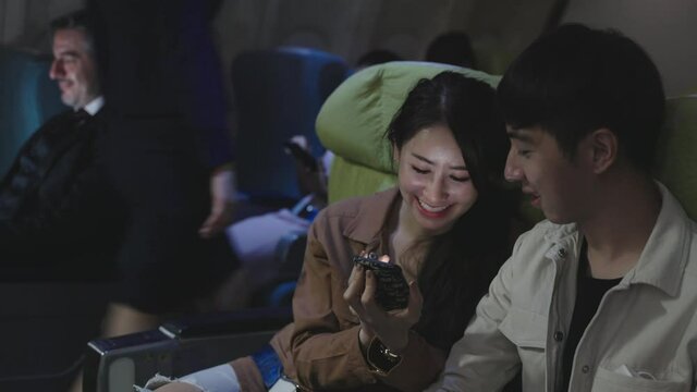Young Asian couple using smartphone together happily while seating in business class during traveling by plane at night. Transportation or technology lifestyle concept.	