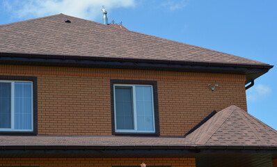 A close-up of a house with an asphalt shingled roofing construction, a roof ventilation cap, plastic soffit, rain gutters, and security cameras installed.