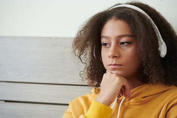 Sad pensive teenage mixed race girl with curly hair listening to music in white headphones and...