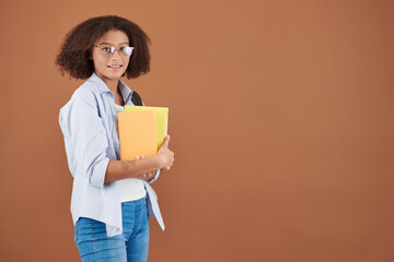 Obraz premium Portrait of smiling mixed-race teenage girl in eyeglasses standing with yellow books against brown background