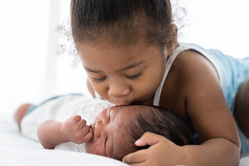 Cute African American little girl kissing on newborn baby cheek on white bed at home. Little girl...