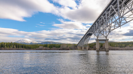 Johnsons Crossing, Teslin River steel Bridge on the Alaska Highway during spring summer time with cloudy, blue sky and magnificent, huge structure over the flowing water below. 