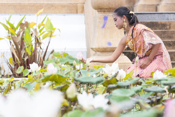 A long-haired Asian girl wearing a traditional Thai dress is reaching out and touching a white lotus in the pool.