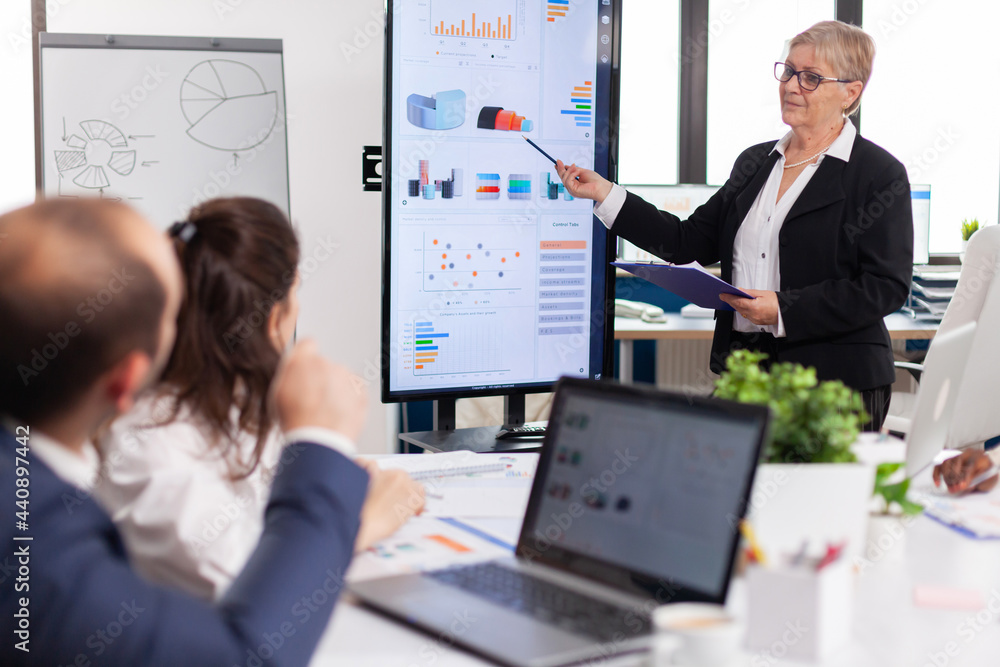 Poster elderly project manager pointing at desktop presenting statistical data, briefing diverse group of e