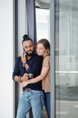 Pretty young woman hugging her husband drinking tea and talking on phone with coworker or employee