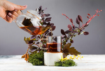 Tea is poured from a teapot into a mug that stands on a podium surrounded by wild plants and moss....