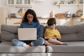 Concentrated mother in eyeglasses and son using laptop and tablet digital computers sitting on couch in living room. Serious mom and little kid work and learn online from home during covid-19 lockdown